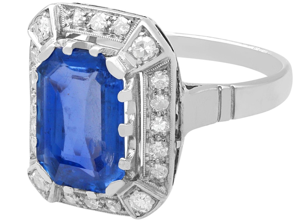 An Art Deco 18ct white gold ring featuring a central octagonal step-cut blue Ceylon sapphire surrounded by a geometric diamond halo with milgrain details. View VI