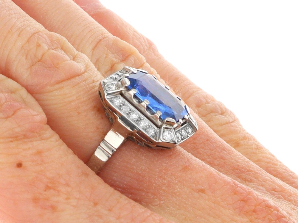 An Art Deco 18ct white gold ring featuring a central octagonal step-cut blue Ceylon sapphire surrounded by a geometric diamond halo with milgrain details. View X