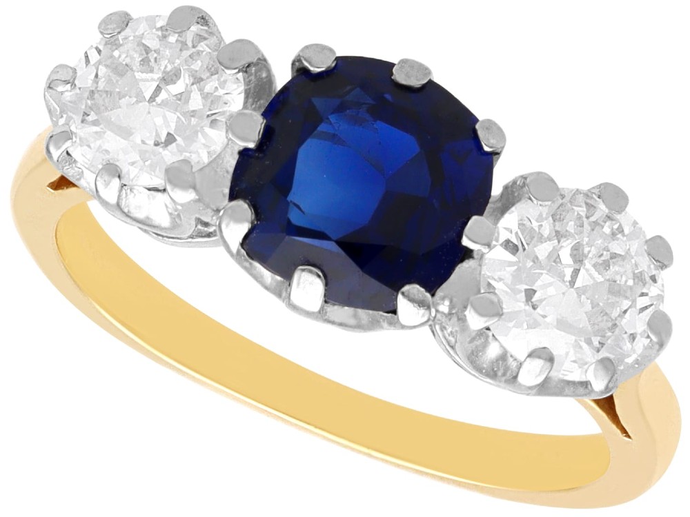 An antique 18ct yellow gold trilogy ring featuring a cushion-shaped blue sapphire flanked by two brilliant-cut diamonds in claw settings.