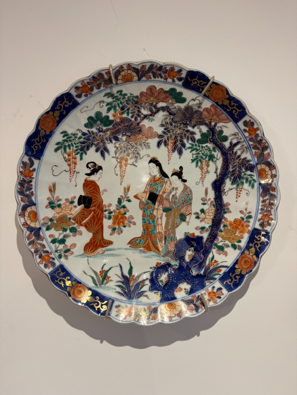A large Japanese Meiji period porcelain charger plate with a scalloped edge, depicting women in traditional kimonos beneath a tree, bordered in cobalt blue and floral motifs.