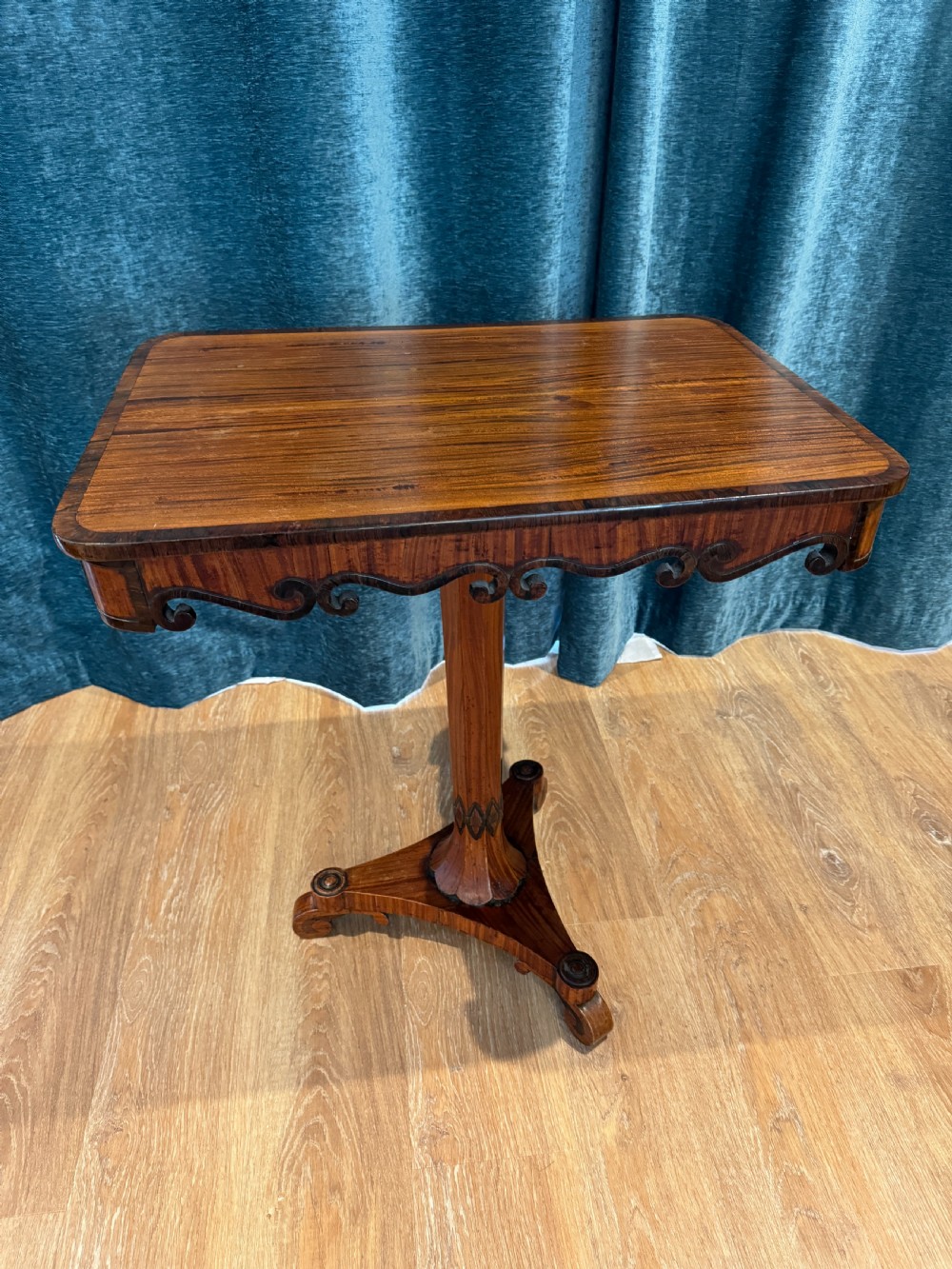 A Regency rosewood side table featuring a crossbanded rectangular top, a carved scrolled apron, and a central pedestal column supported by a triform base on scroll feet.