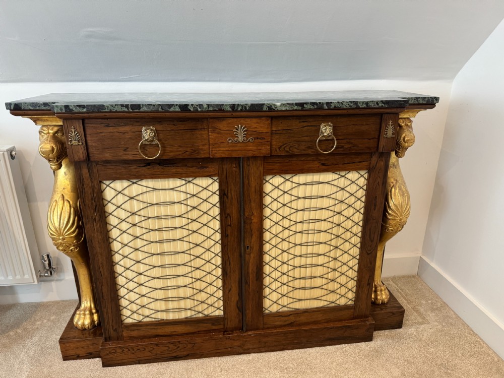 A Regency rosewood side cabinet featuring a dark green marble top, gilt lion monopodia legs, and two brass grille doors lined with pleated fabric.