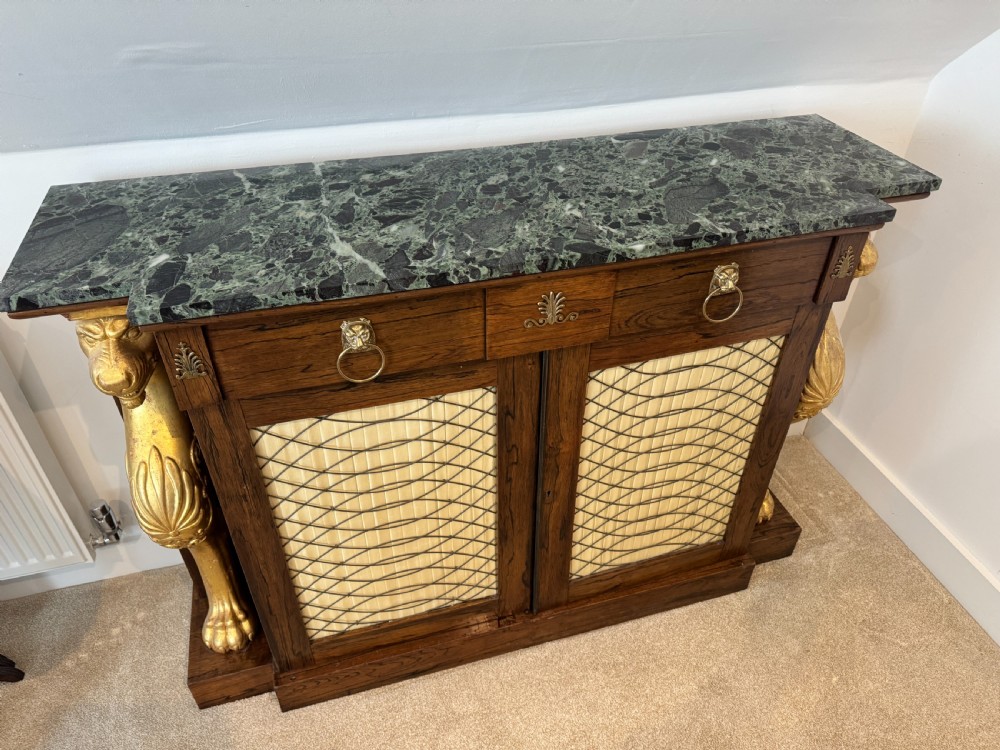 A Regency rosewood side cabinet featuring a dark green marble top, gilt lion monopodia legs, and two brass grille doors lined with pleated fabric. View II