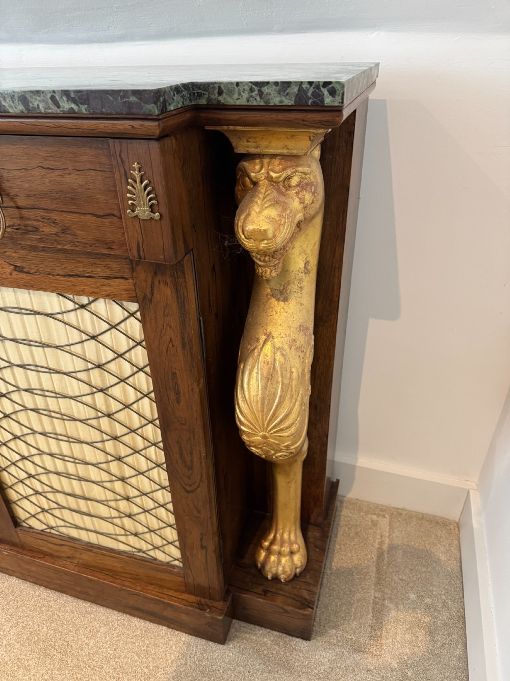 A Regency rosewood side cabinet featuring a dark green marble top, gilt lion monopodia legs, and two brass grille doors lined with pleated fabric. View VI