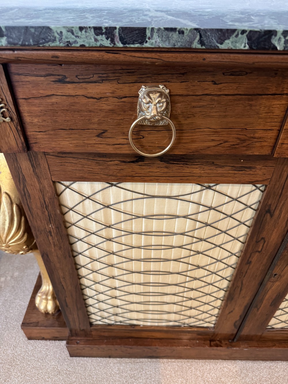 A Regency rosewood side cabinet featuring a dark green marble top, gilt lion monopodia legs, and two brass grille doors lined with pleated fabric. View VII