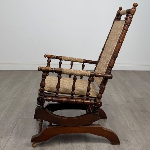 fig 1. An American 20th-century platform rocking chair crafted from dark-stained beech with turned spindle arms, patterned gold upholstery, and a spring-loaded base on brass castors.