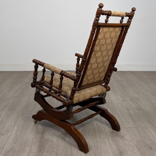 fig 2. An American 20th-century platform rocking chair crafted from dark-stained beech with turned spindle arms, patterned gold upholstery, and a spring-loaded base on brass castors.