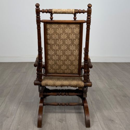 fig 3. An American 20th-century platform rocking chair crafted from dark-stained beech with turned spindle arms, patterned gold upholstery, and a spring-loaded base on brass castors.