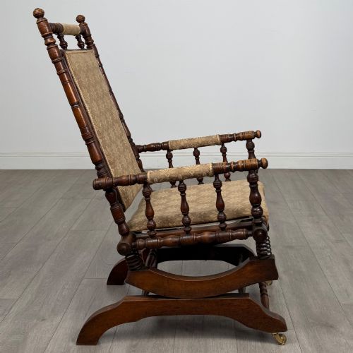 fig 5. An American 20th-century platform rocking chair crafted from dark-stained beech with turned spindle arms, patterned gold upholstery, and a spring-loaded base on brass castors.