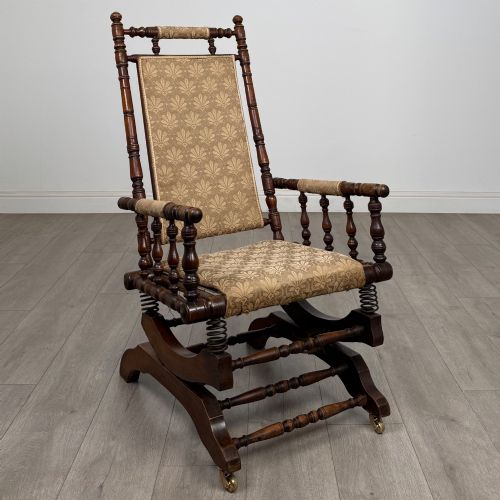 fig 6. An American 20th-century platform rocking chair crafted from dark-stained beech with turned spindle arms, patterned gold upholstery, and a spring-loaded base on brass castors.