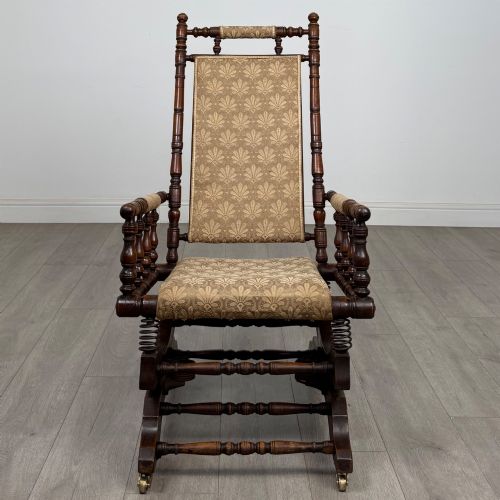 fig 7. An American 20th-century platform rocking chair crafted from dark-stained beech with turned spindle arms, patterned gold upholstery, and a spring-loaded base on brass castors.
