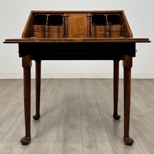 fig 1. A golden oak slant-front writing bureau standing on four slender legs with pad feet, featuring a single lower drawer with drop handles and a hinged writing slope.