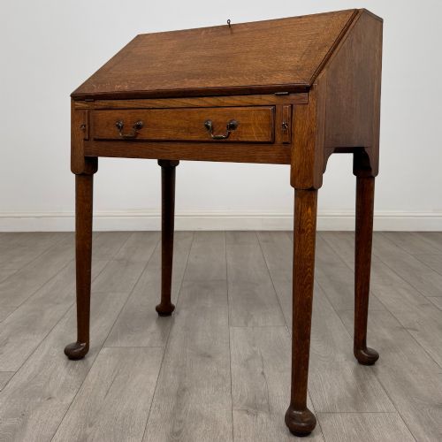 fig 2. A golden oak slant-front writing bureau standing on four slender legs with pad feet, featuring a single lower drawer with drop handles and a hinged writing slope.