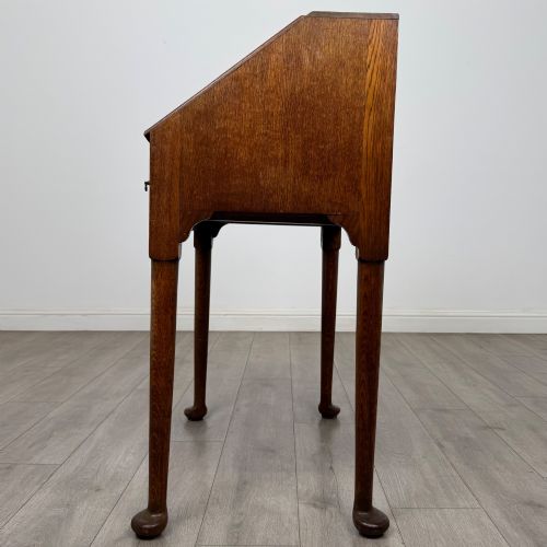 fig 3. A golden oak slant-front writing bureau standing on four slender legs with pad feet, featuring a single lower drawer with drop handles and a hinged writing slope.