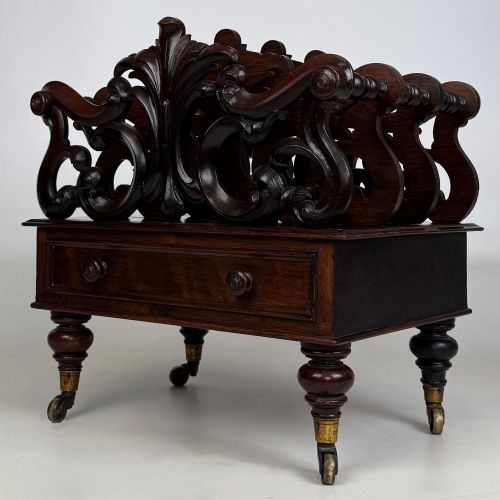 fig 1. A dark rosewood Victorian canterbury with central acanthus carving, double scrollwork dividers, a single lower drawer, turned legs, and brass castors.