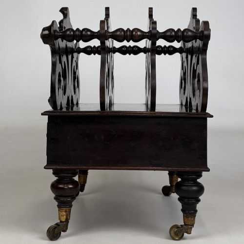 fig 2. A dark rosewood Victorian canterbury with central acanthus carving, double scrollwork dividers, a single lower drawer, turned legs, and brass castors.