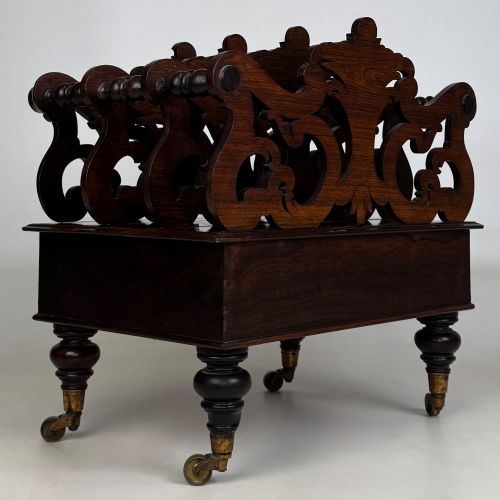 fig 3. A dark rosewood Victorian canterbury with central acanthus carving, double scrollwork dividers, a single lower drawer, turned legs, and brass castors.
