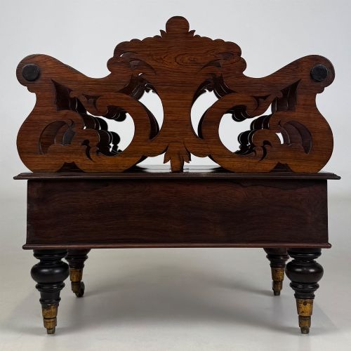 fig 4. A dark rosewood Victorian canterbury with central acanthus carving, double scrollwork dividers, a single lower drawer, turned legs, and brass castors.