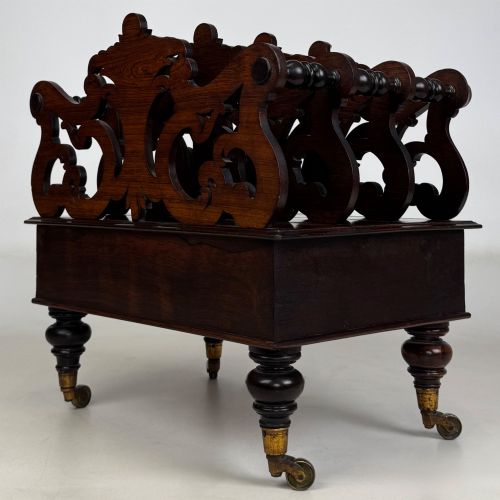 fig 5. A dark rosewood Victorian canterbury with central acanthus carving, double scrollwork dividers, a single lower drawer, turned legs, and brass castors.