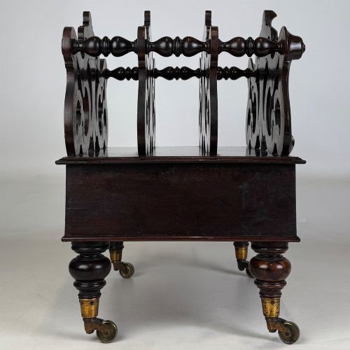 fig 6. A dark rosewood Victorian canterbury with central acanthus carving, double scrollwork dividers, a single lower drawer, turned legs, and brass castors.