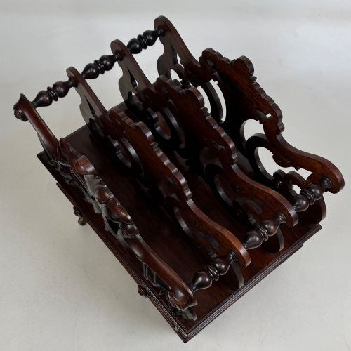 fig 8. A dark rosewood Victorian canterbury with central acanthus carving, double scrollwork dividers, a single lower drawer, turned legs, and brass castors.