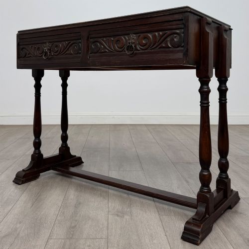fig 1. A dark brown carved oak side table featuring two scroll-carved front drawers with metal drop handles and turned trestle legs.