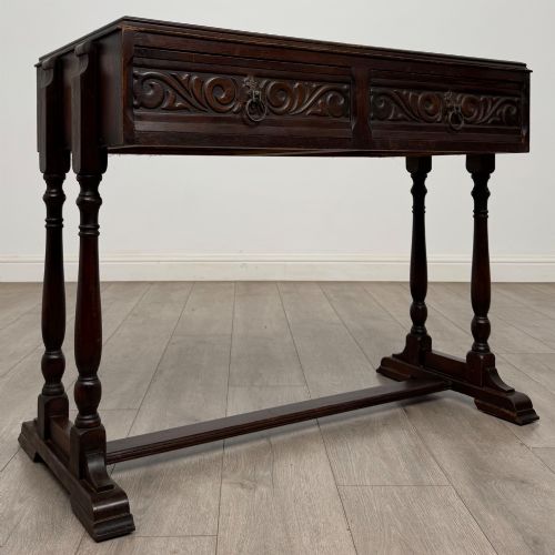 fig 7. A dark brown carved oak side table featuring two scroll-carved front drawers with metal drop handles and turned trestle legs.