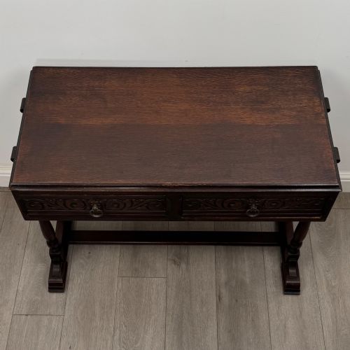 fig 8. A dark brown carved oak side table featuring two scroll-carved front drawers with metal drop handles and turned trestle legs.