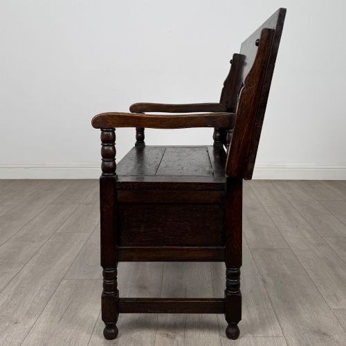 fig 3. A dark oak monks bench featuring a panelled backrest with a central diamond motif, turned arm supports, and a lift-up seat.