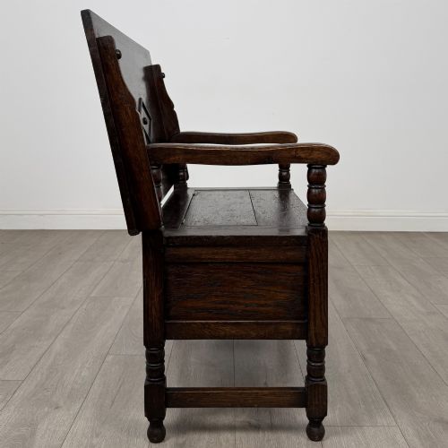 fig 7. A dark oak monks bench featuring a panelled backrest with a central diamond motif, turned arm supports, and a lift-up seat.