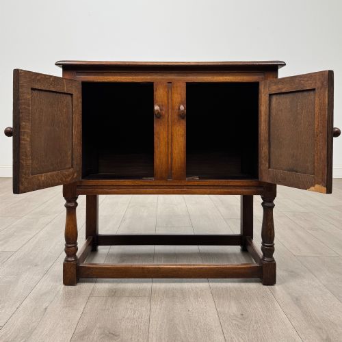 fig 1. A small warm brown carved oak cupboard with double doors featuring carved arches, iron hinges, turned legs, and a lower stretcher base.