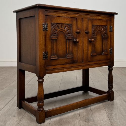 fig 8. A small warm brown carved oak cupboard with double doors featuring carved arches, iron hinges, turned legs, and a lower stretcher base.