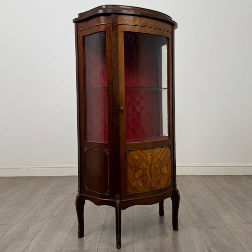 fig 8. A French mahogany display vitrine featuring a curved glass door, red fabric interior lining, floral marquetry lower panel, and brass leg mounts.