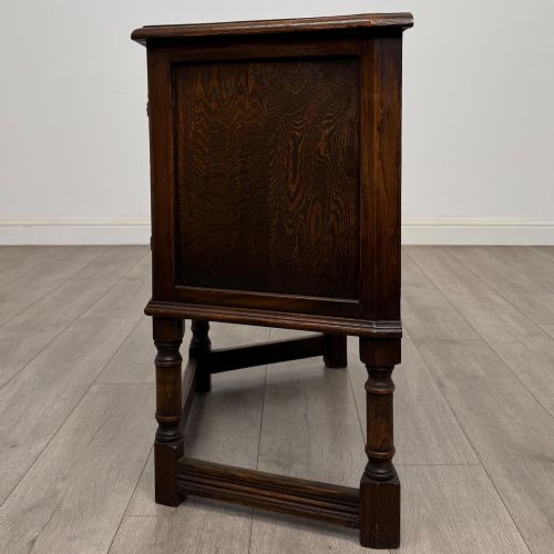 fig 3. A small dark oak cupboard featuring a single carved panel door, turned legs, a lower stretcher, and decorative brass hinges.