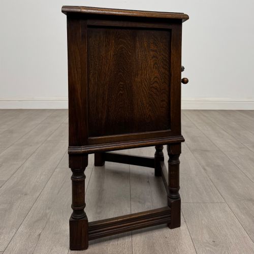 fig 7. A small dark oak cupboard featuring a single carved panel door, turned legs, a lower stretcher, and decorative brass hinges.