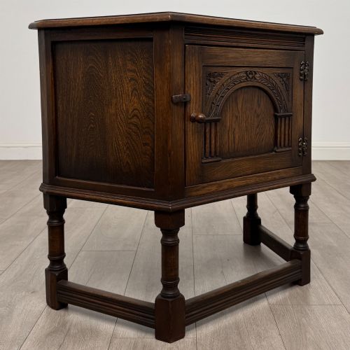 fig 8. A small dark oak cupboard featuring a single carved panel door, turned legs, a lower stretcher, and decorative brass hinges.