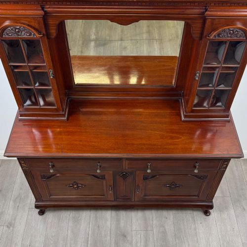 fig 9. An Arts and Crafts dark mahogany sideboard featuring an upper mirrored superstructure flanked by leaded glass cabinet doors above lower drawers and carved cupboards.