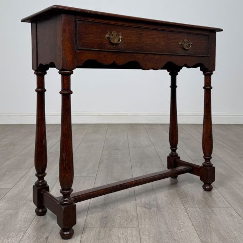 fig 7. A dark oak side table featuring a single wide drawer with brass drop handles, a scalloped apron, turned legs, and lower stretchers.