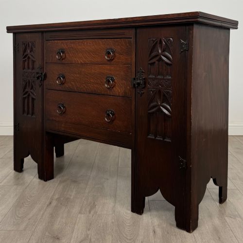 fig 2. A dark oak Gothic Revival sideboard featuring three central drawers with metal drop ring handles, flanked by two carved tracery panel doors with decorative iron latches.