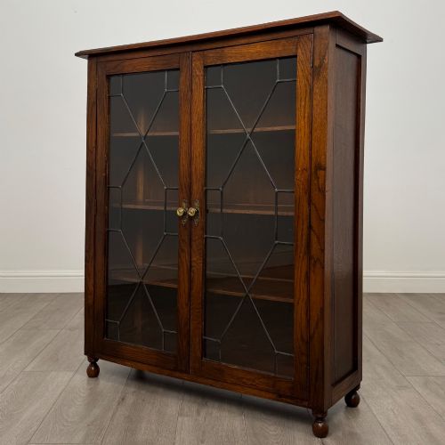 fig 2. An Edwardian warm brown oak display cabinet featuring twin leaded glass doors with a geometric pattern, interior wooden shelves, and rounded bun feet.