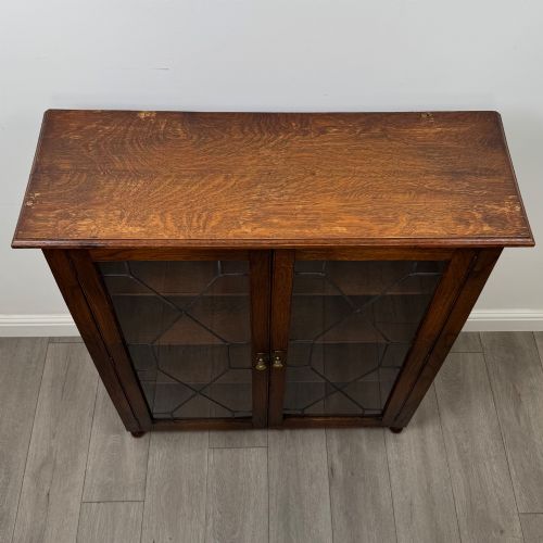 fig 9. An Edwardian warm brown oak display cabinet featuring twin leaded glass doors with a geometric pattern, interior wooden shelves, and rounded bun feet.