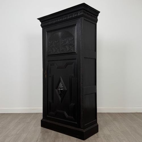 fig 2. A single-door dark oak Victorian wardrobe featuring geometric panel mouldings, a carved relief face in a central diamond, and a pronounced decorative cornice.