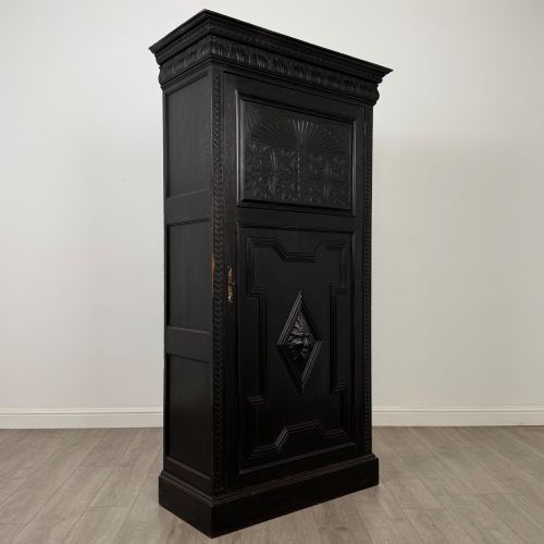 fig 8. A single-door dark oak Victorian wardrobe featuring geometric panel mouldings, a carved relief face in a central diamond, and a pronounced decorative cornice.