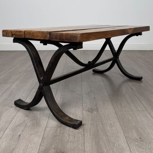 fig 2. A low rectangular coffee table featuring a thick hardwood plank top set upon a dark curved iron X-frame base.