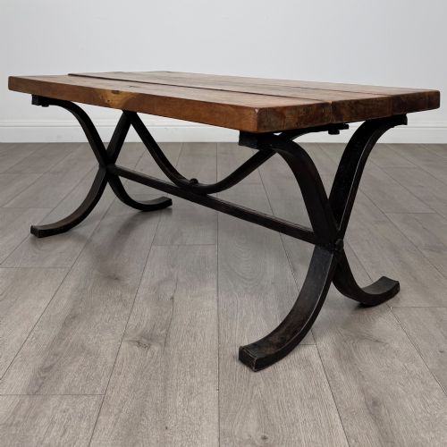 fig 4. A low rectangular coffee table featuring a thick hardwood plank top set upon a dark curved iron X-frame base.