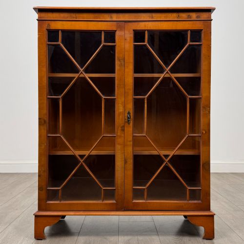 fig 1. An antique yew wood display cabinet featuring two astragal glazed doors with geometric glazing bars, housing books on internal shelves, and resting on bracket feet.