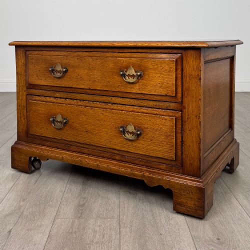fig 2. A low two-drawer oak chest featuring raised panel fronts, decorative brass drop handles, and bracket feet in a warm brown finish.