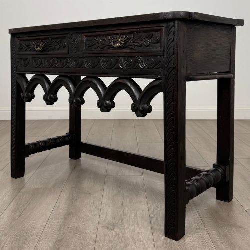fig 1. A dark oak 19th-century side table featuring floral relief carvings on two drawer fronts, a row of carved cusped arches along the lower apron, and turned side stretchers.