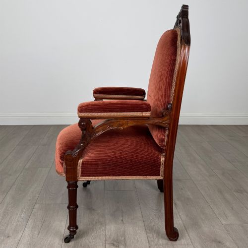 fig 1. A Victorian rosewood armchair featuring a dark timber frame, carved scroll detailing, rust red velvet upholstery, and turned front legs on castors.