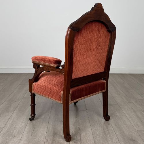 fig 2. A Victorian rosewood armchair featuring a dark timber frame, carved scroll detailing, rust red velvet upholstery, and turned front legs on castors.